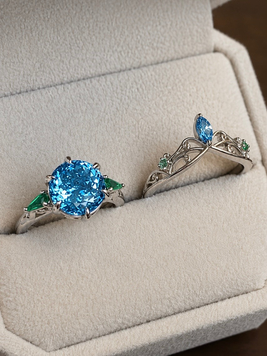 Platinum engagement ring with a blue lab-grown diamond and emerald accents beside a tiara-inspired wedding band